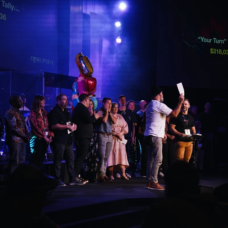 A group of people on a stage reacts with surprise and joy as a man announces a fundraising total of $318,036, displayed on a screen behind them.
